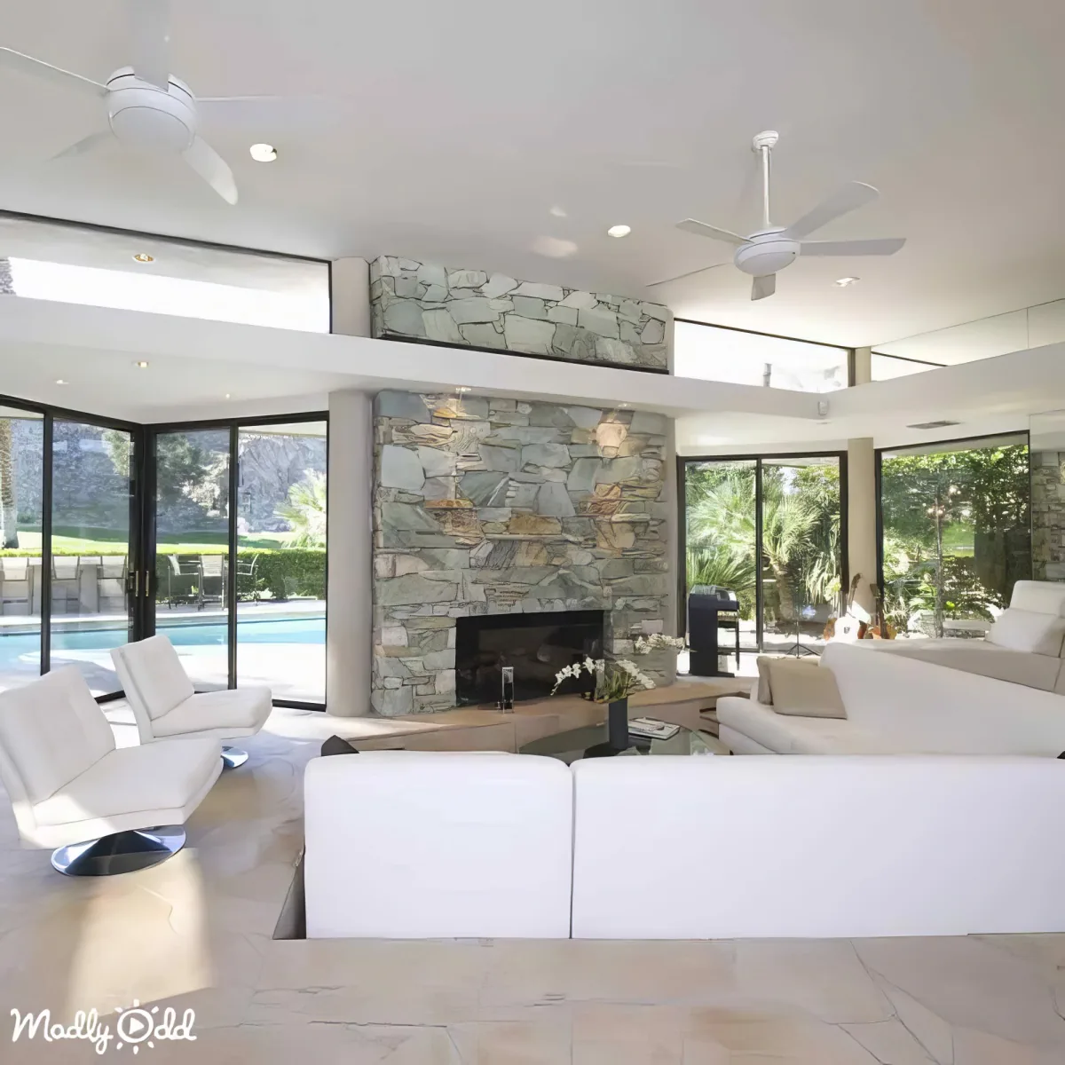 A modern living room with white furniture, a stone fireplace, and large windows overlooking a pool and greenery.