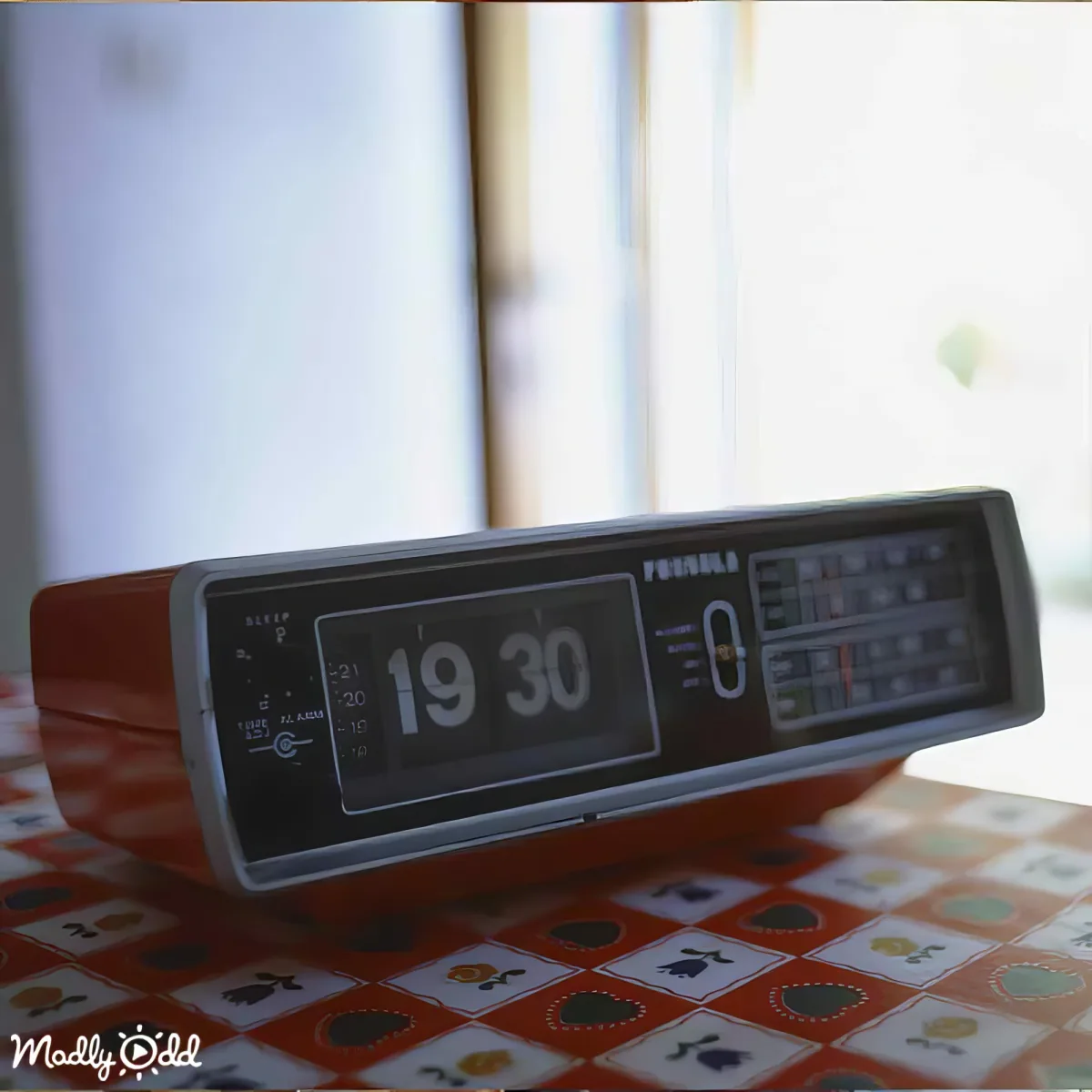 A retro flip clock displaying 19:30 sits on a patterned tablecloth near a window.