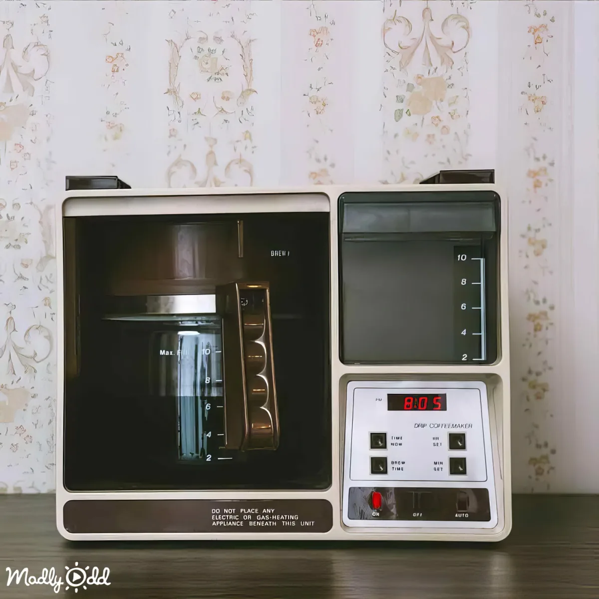 A retro-style drip coffee maker with a digital display and control panel sits on a table against floral wallpaper.