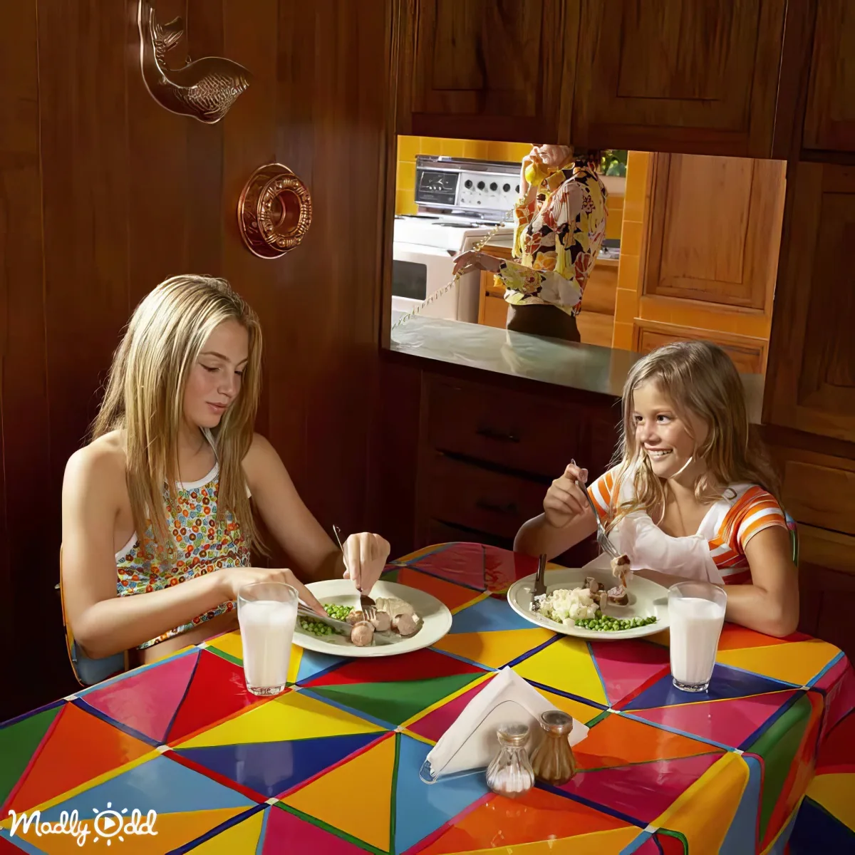 Two girls sit at a colorful table, eating, while a person in the kitchen holds a phone to their ear.