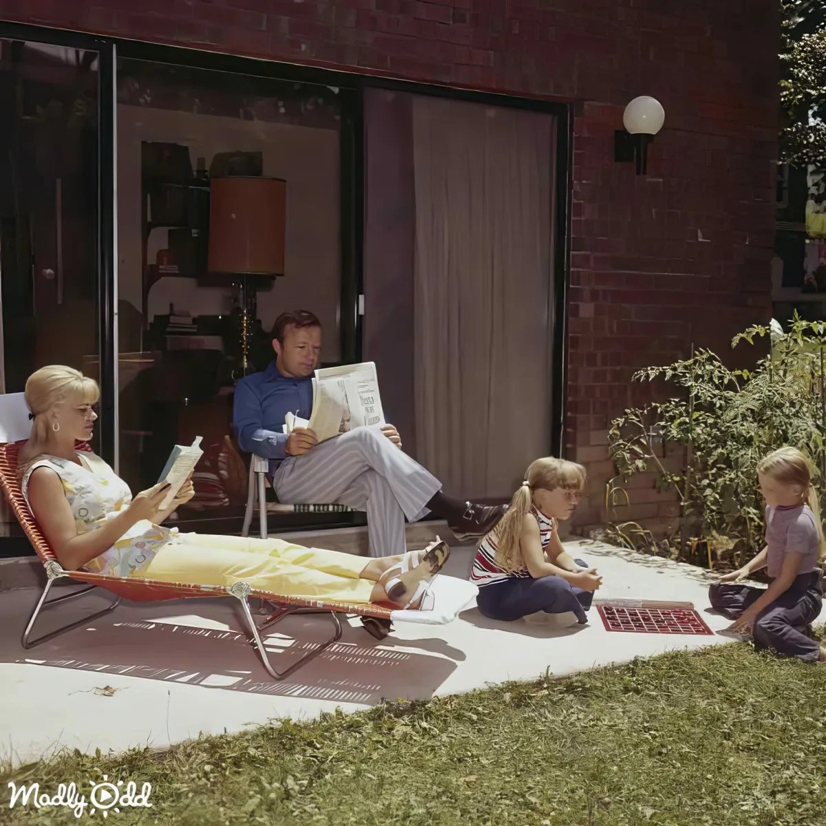 A woman lounges on a chair, reading, while a man and two children sit nearby, enjoying a sunny day outside a brick house.