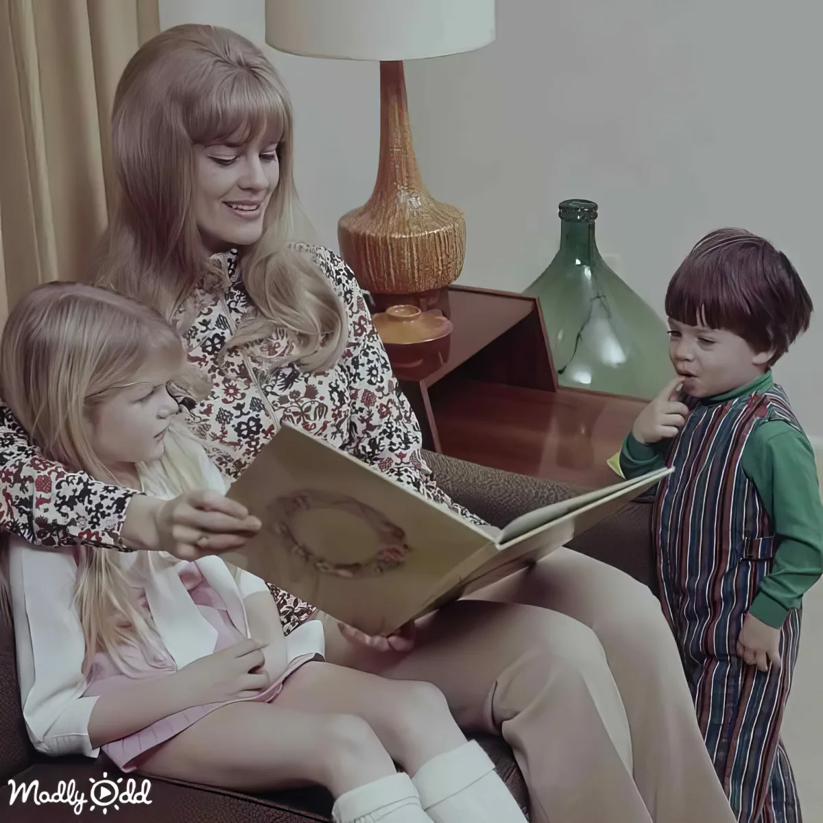A woman reads a book to two children in a cozy, retro-style living room.