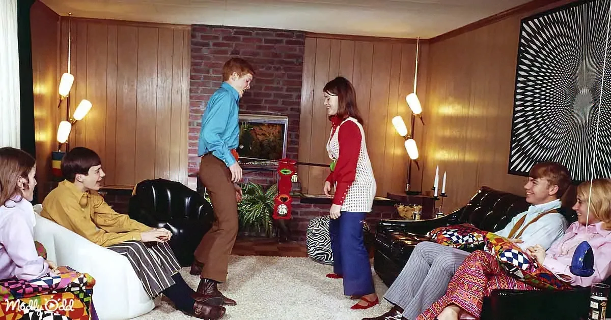 A woman in a red sweater stands in a retro living room, talking to a man in a blue shirt, while others sit on couches and...