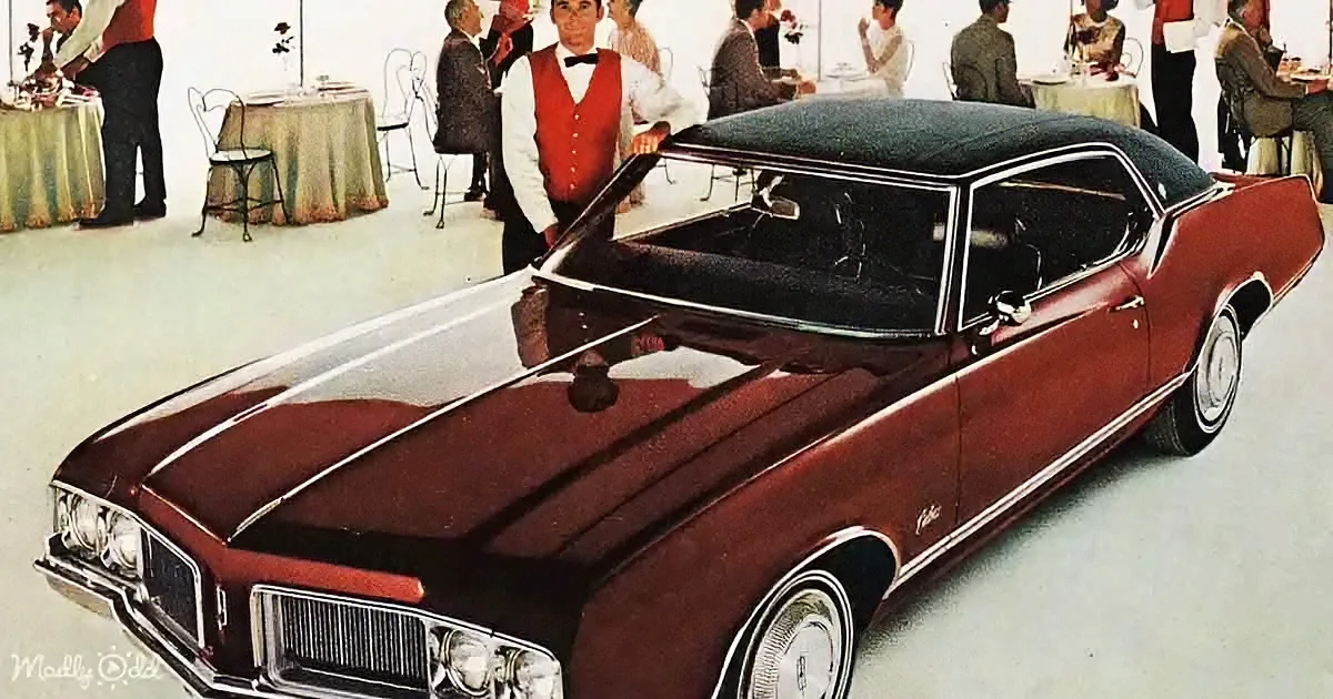 A maroon vintage car with white trim is displayed in a showroom with people in the background.