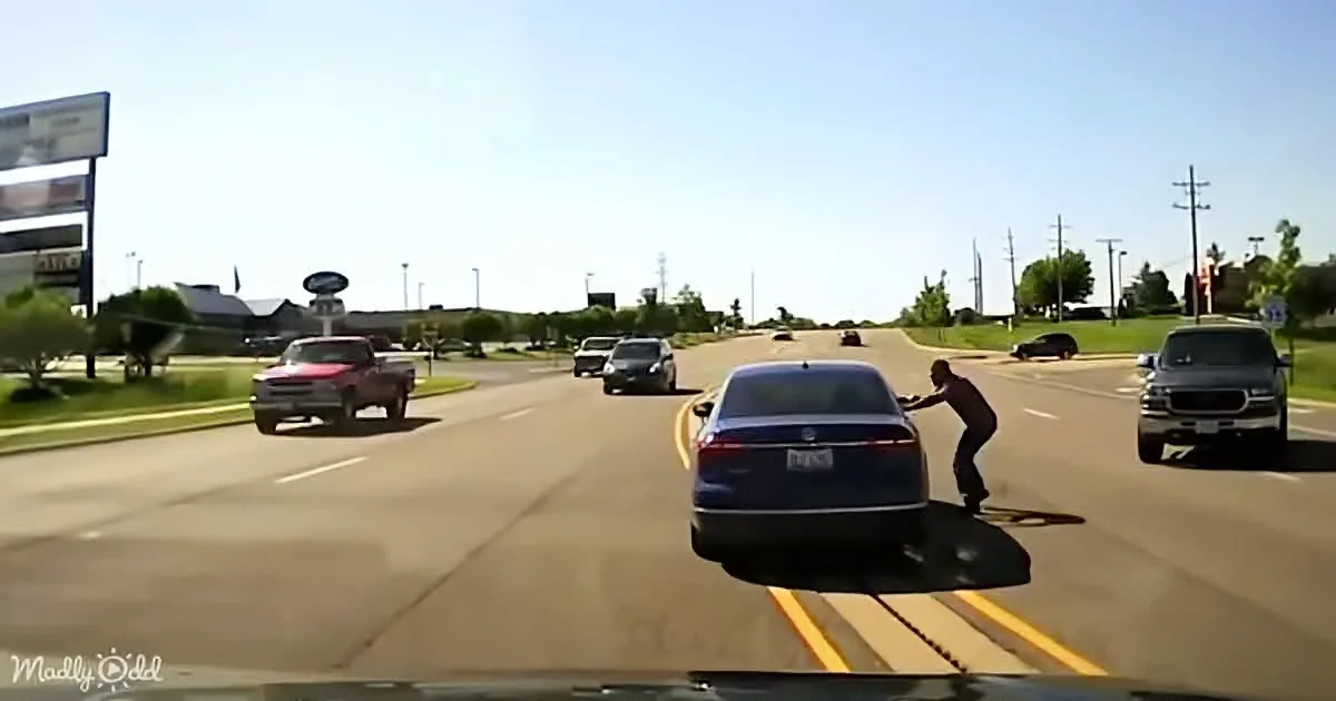 A man is pushing a car on a road, with other vehicles and a clear sky in the background.