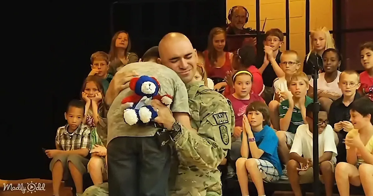 A soldier in uniform hugs a child holding a patriotic teddy bear, surrounded by a group of children watching.