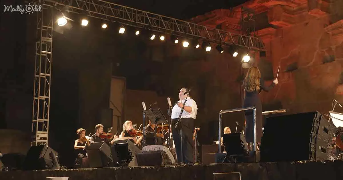 A conductor leads an orchestra on stage with a backdrop of red rock formations.