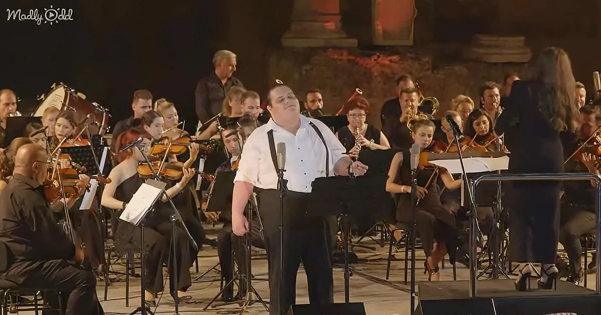A man in white shirt and black pants sings into a microphone, surrounded by an orchestra.
