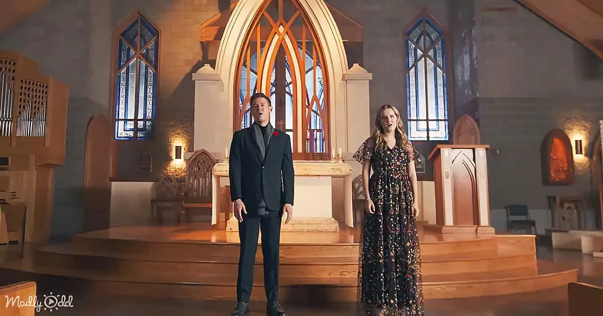 A man in a suit and a woman in a floral dress stand on a church stage, surrounded by stained glass windows and wooden...