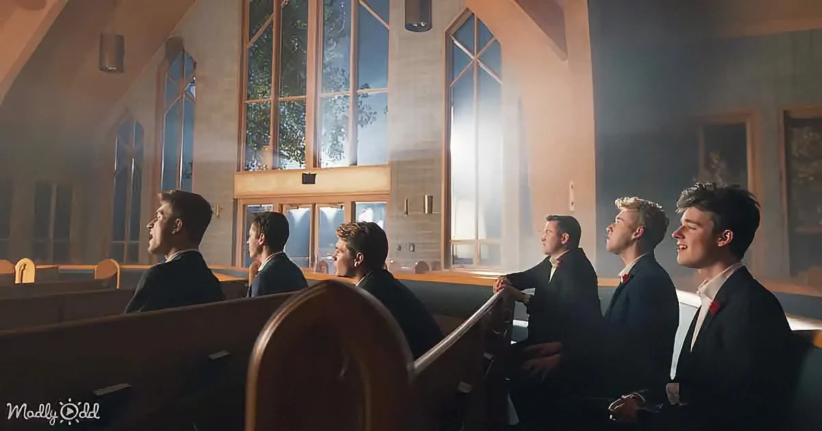 Six young men in suits sing in a church with stained glass windows and wooden pews.