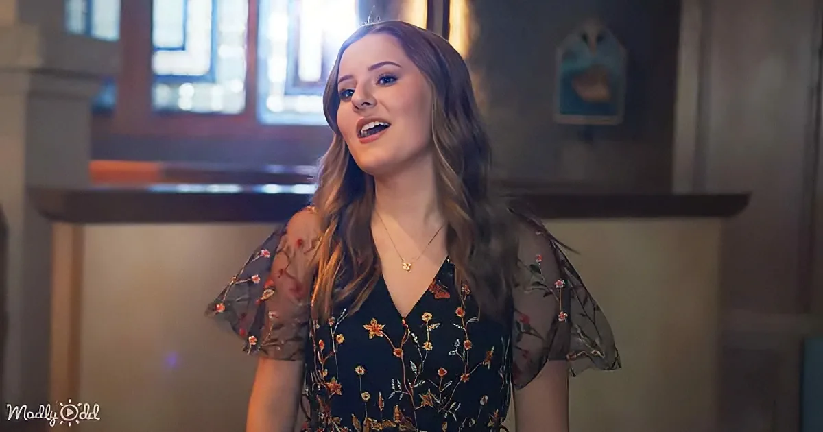 A woman with long brown hair, wearing a floral dress, stands indoors with a surprised expression.