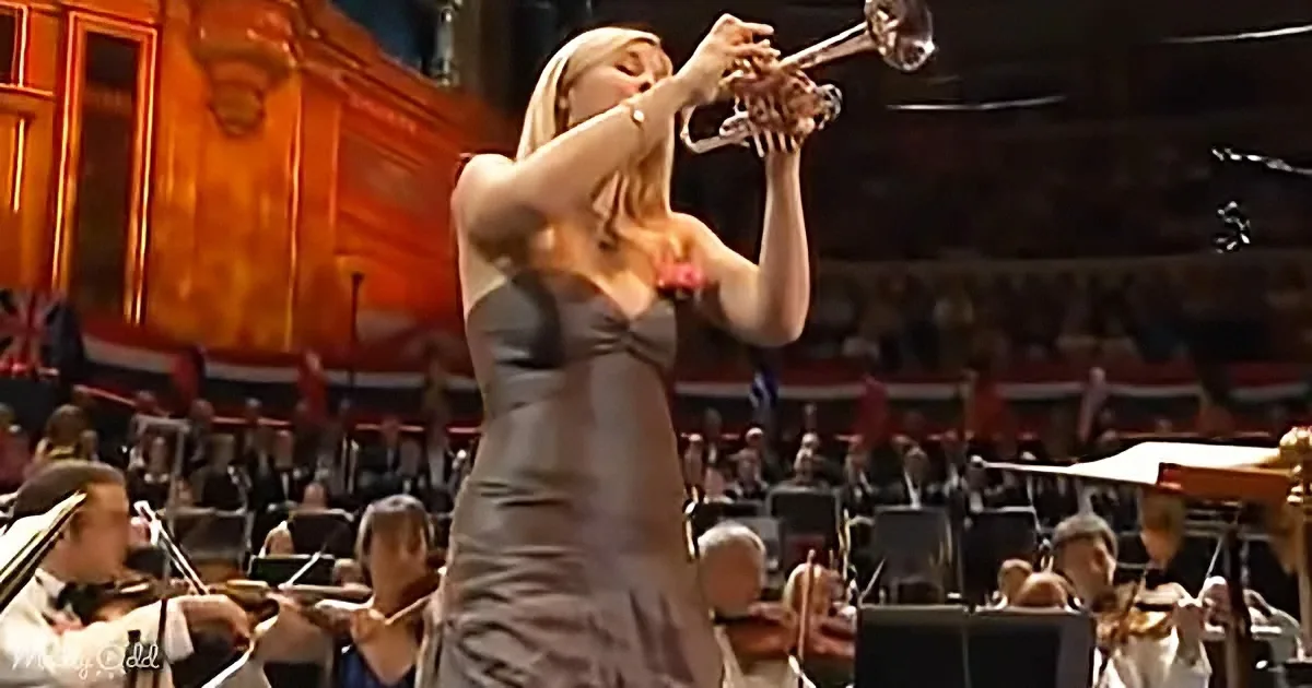 A woman in a black dress plays trumpet on stage, surrounded by an orchestra in a grand hall.