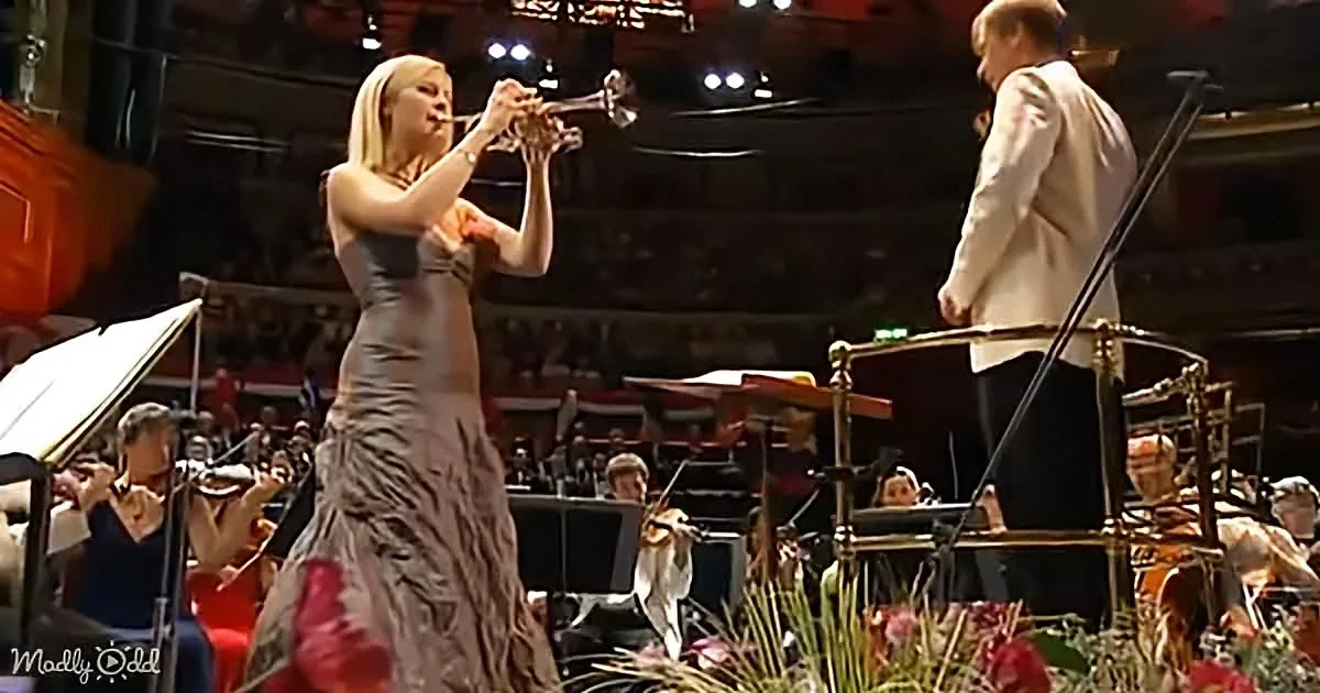 A woman in a long dress plays trumpet on stage, with an orchestra and conductor in the background.
