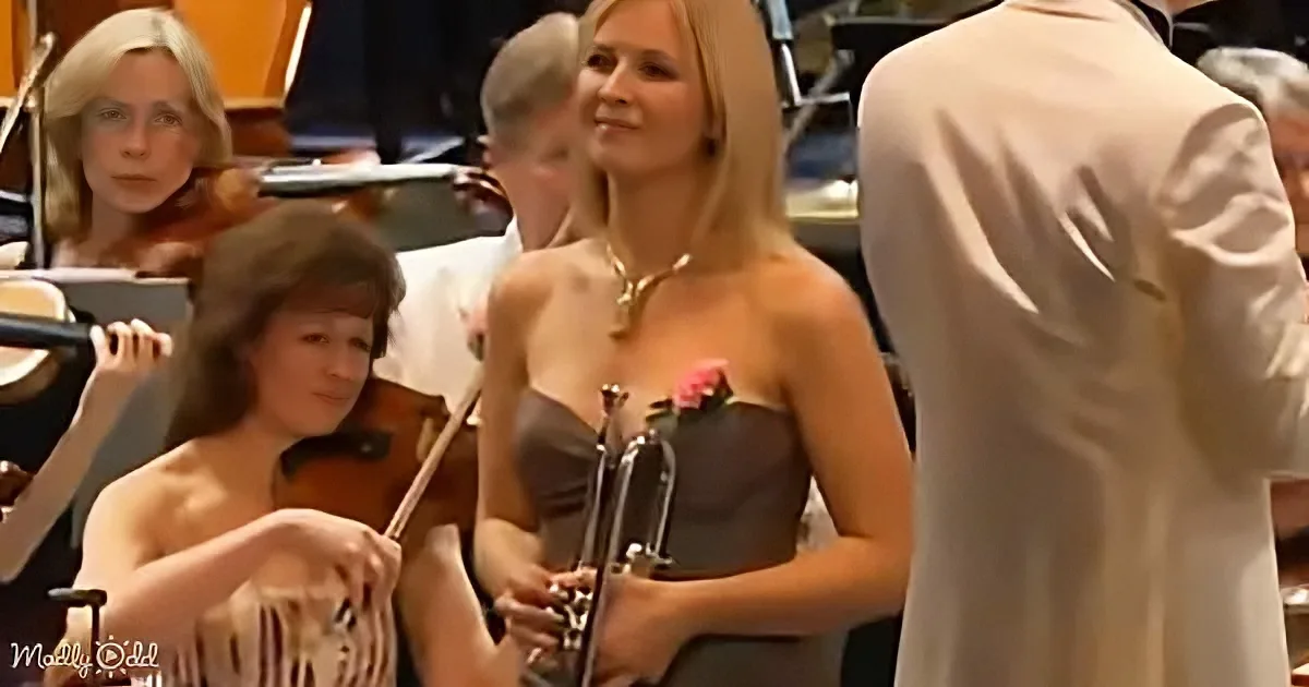 A woman in a strapless dress holds a trumpet, smiling, with musicians in the background.