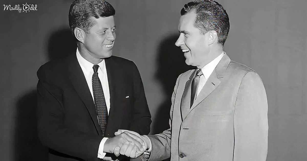 Two men in suits shake hands, both smiling, against a plain background.