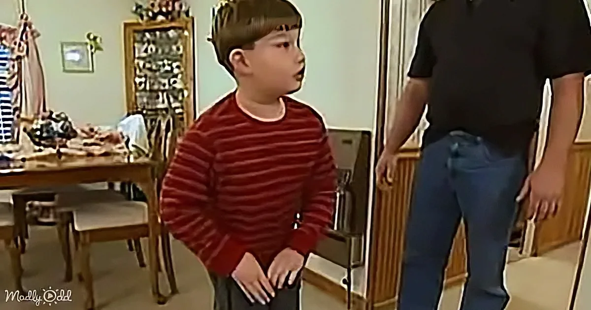 A young boy in a red striped sweater stands in a kitchen, looking up at an adult.