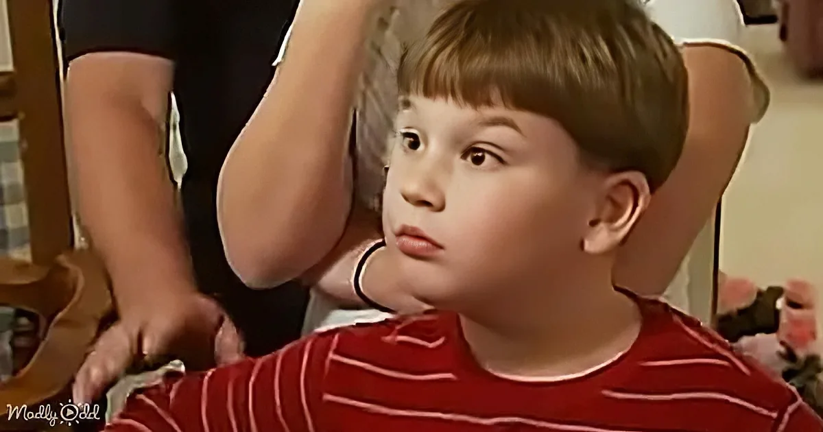 A young boy in a red striped shirt sits with his arm raised, looking to the side.