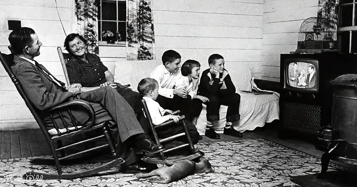 A family gathers around a vintage television, watching a show together on a porch.