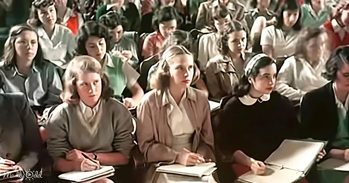 A group of women in vintage attire sit in rows, some with notebooks, in what appears to be a classroom or lecture hall.