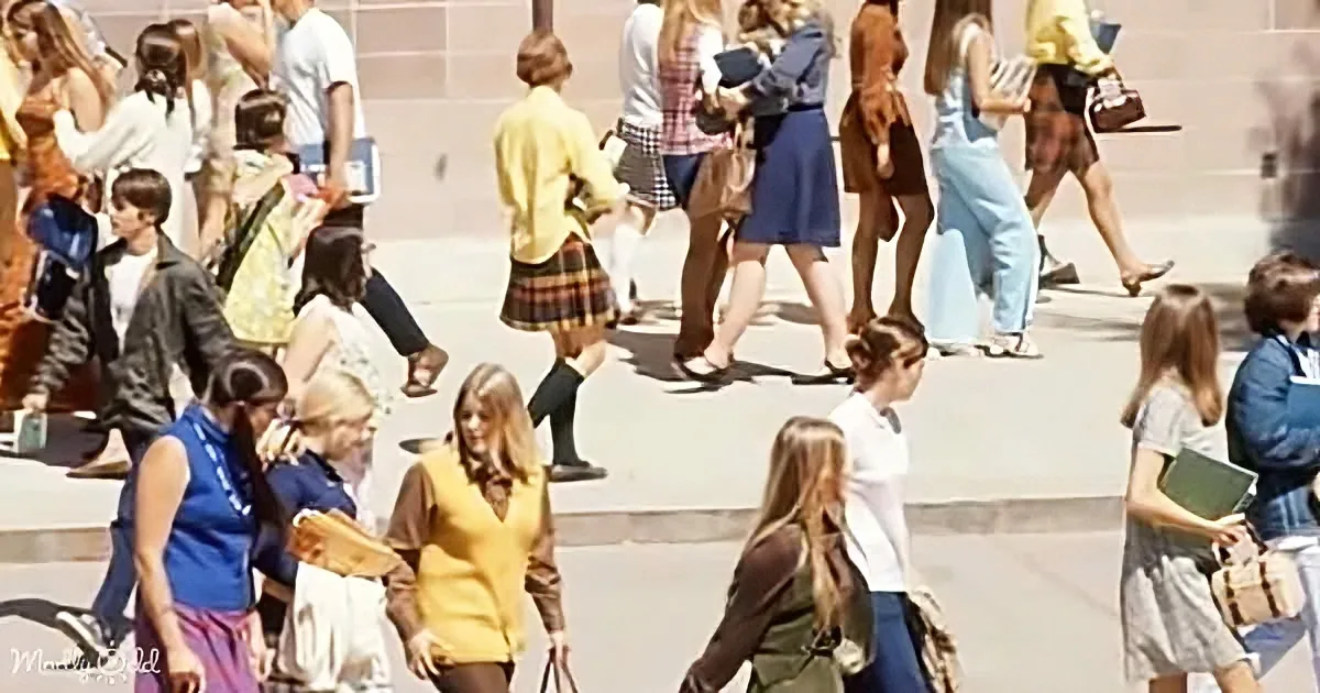 A woman in a yellow sweater and brown pants walks among a crowd of people on a sunny day.