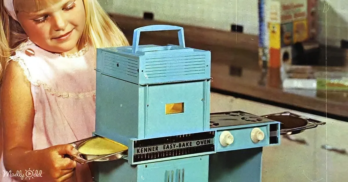 A young girl in a pink dress is holding a baking dish next to a blue Kenner Easy-Bake Oven.