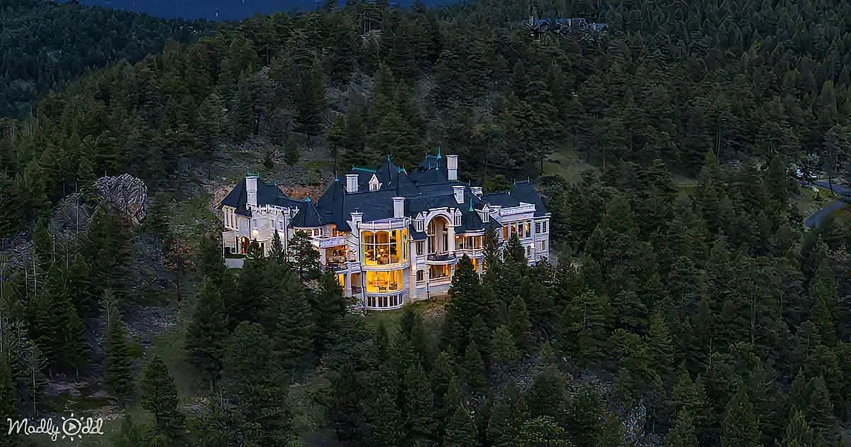 A grand, illuminated mansion sits surrounded by dense evergreen forest at dusk.
