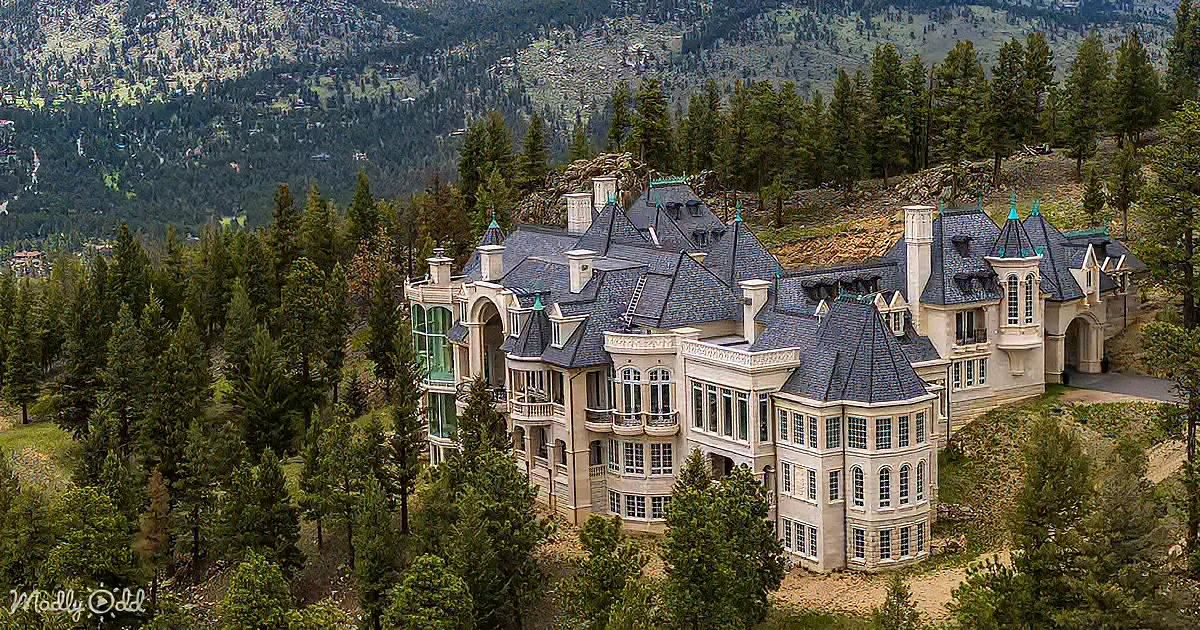 A grand, multi-story mansion with a slate roof sits amidst dense evergreen trees, surrounded by a mountainous landscape.