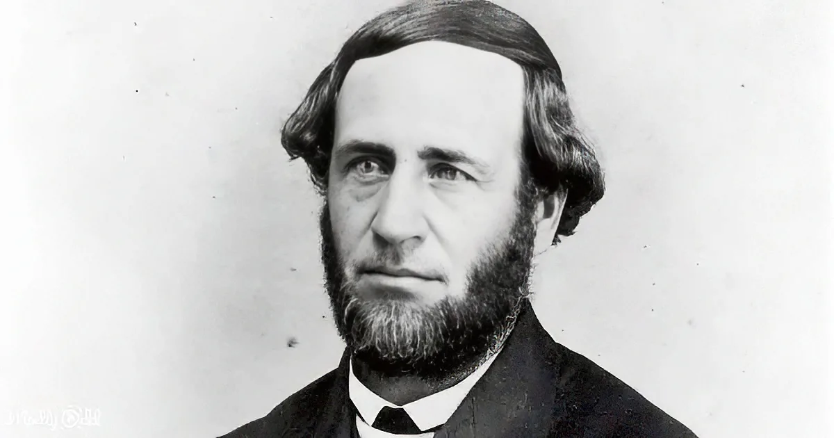 A man with a neatly groomed beard and hair, wearing formal attire, against a plain background.