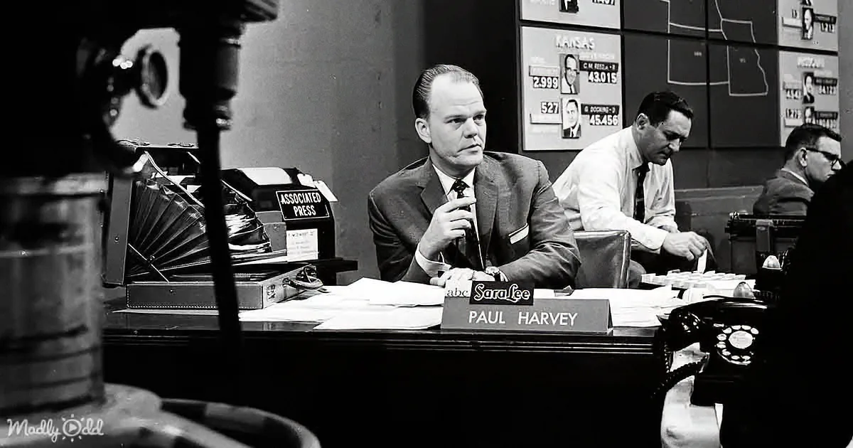 Paul Harvey sits at a desk with a microphone, surrounded by other workers in a newsroom setting.