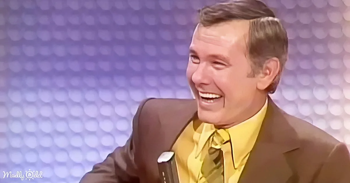 A man in a brown suit and yellow shirt is laughing, seated against a patterned background.