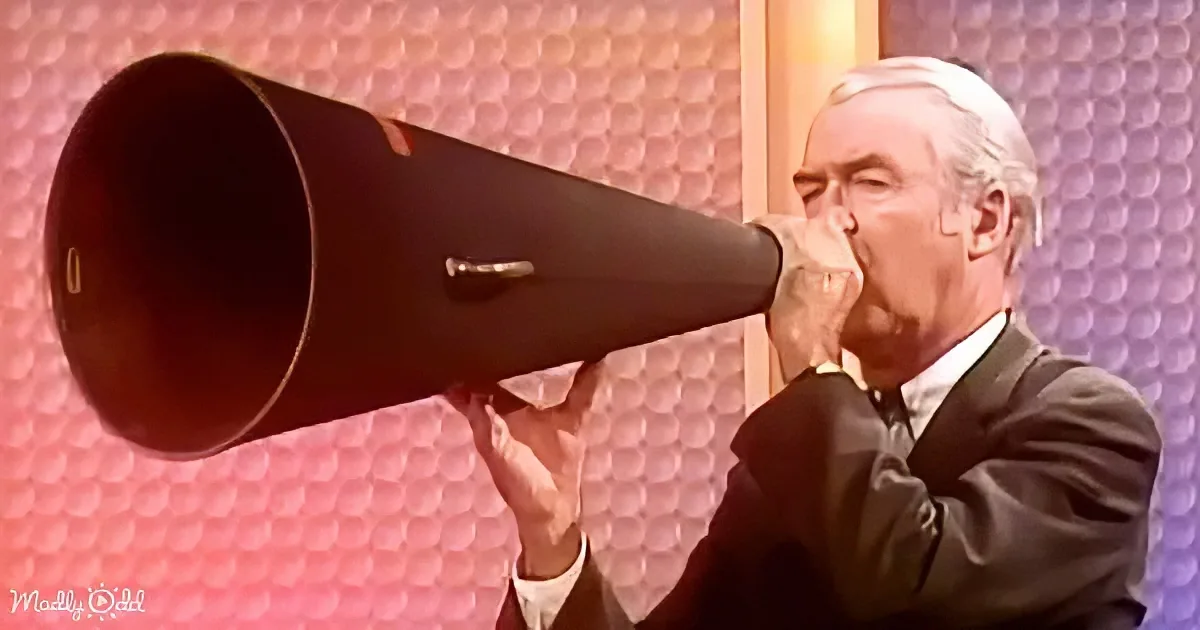 A man in a suit holds a large megaphone to his mouth, seemingly shouting.