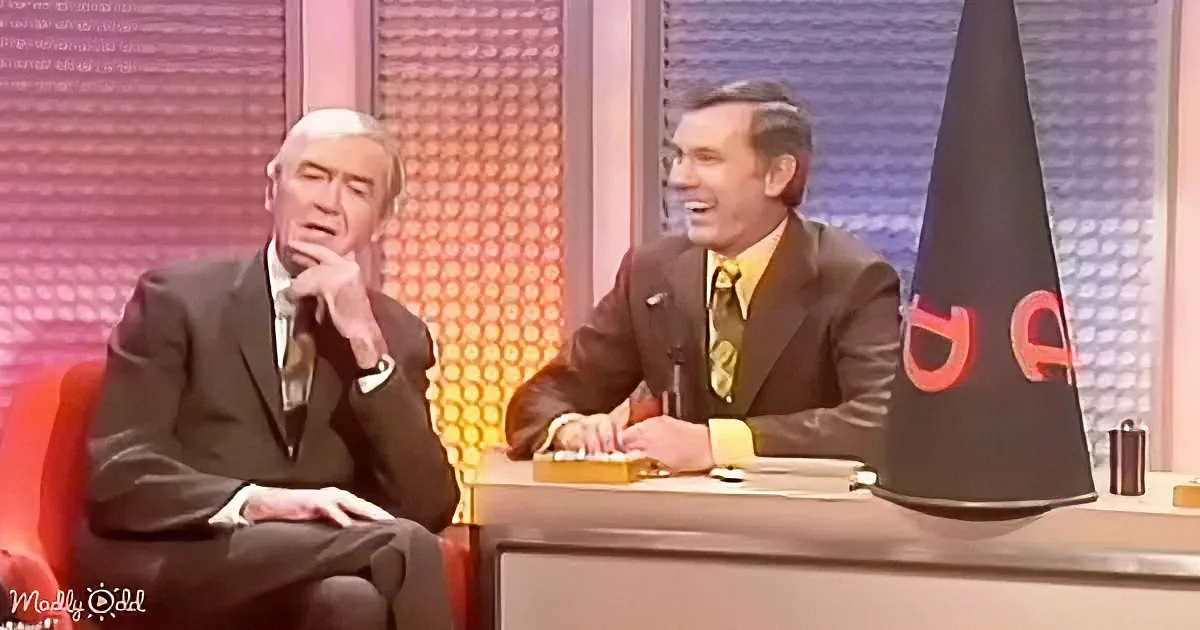 Two men in suits, one seated on a red chair, the other at a desk with a microphone, in a studio setting.