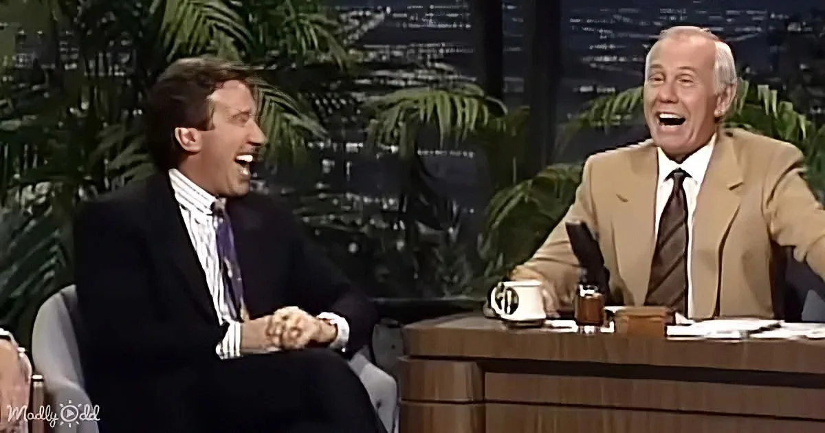 Two men in suits share a laugh on a talk show set with a cityscape backdrop.