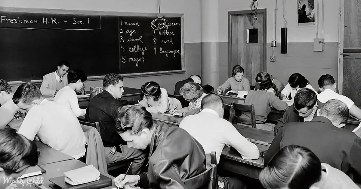 Students sit at desks in a classroom, writing, with a blackboard listing "Freshman H.R. - Sec. 1" and questions.