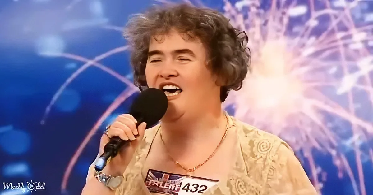 A person with curly hair sings into a microphone on a stage with fireworks in the background.