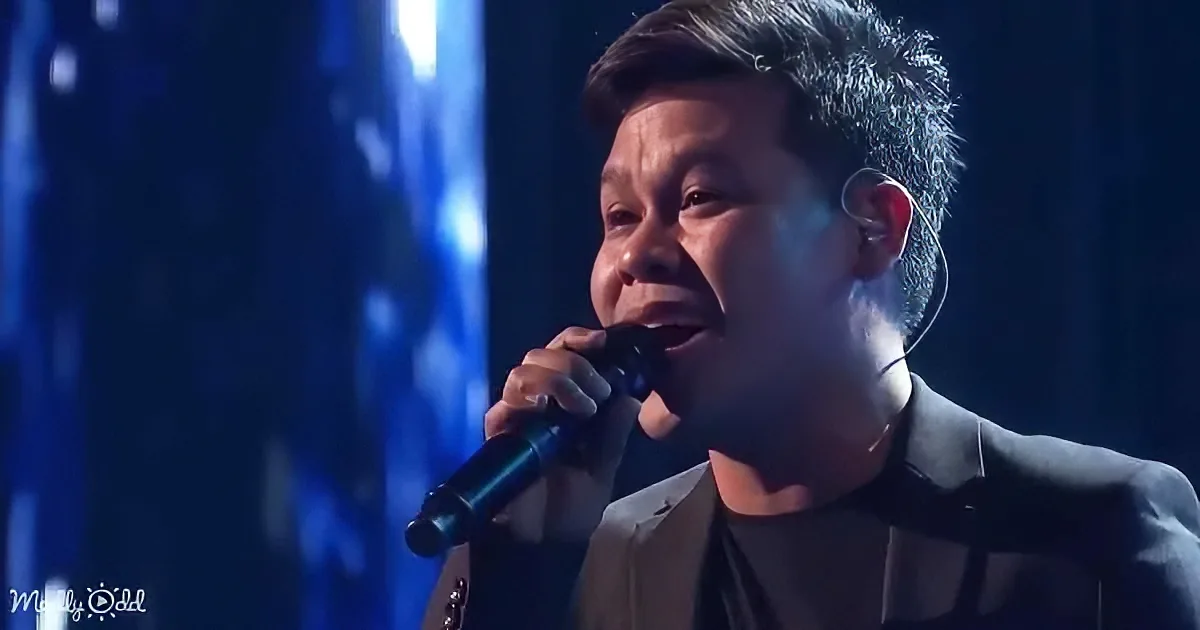 A person in a suit sings into a microphone on stage with blue lighting.