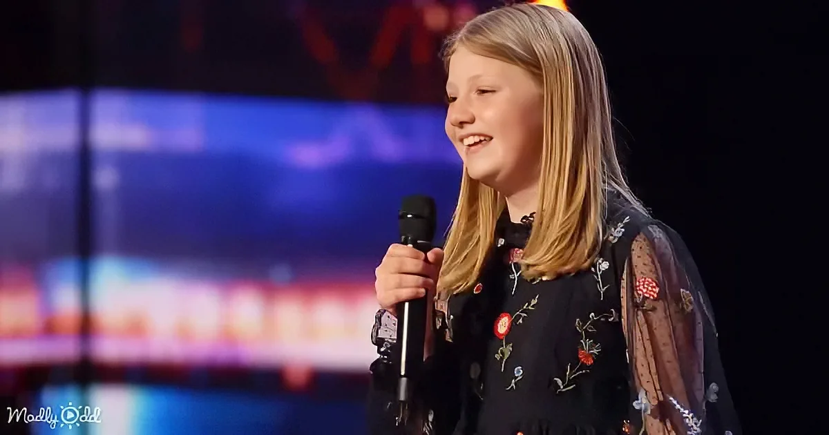 A young girl with long blonde hair smiles while holding a microphone on stage.