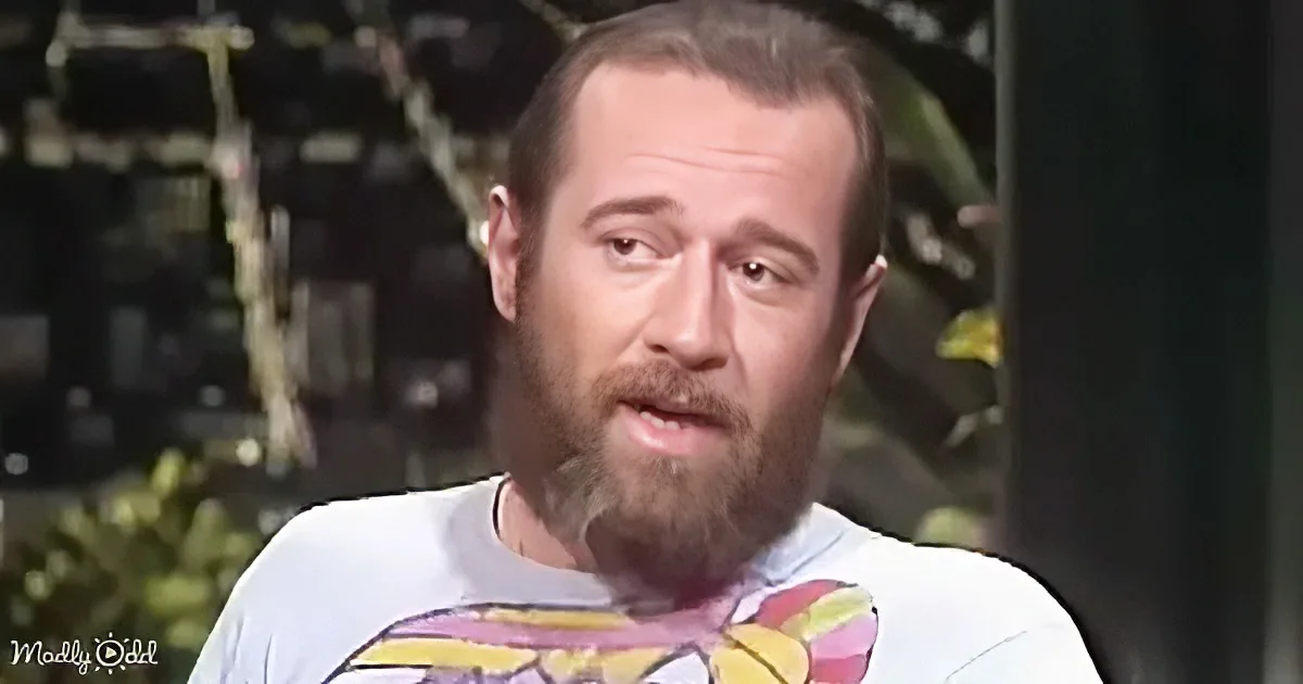 A man with a beard and mustache wearing a white t-shirt with a colorful design is speaking.