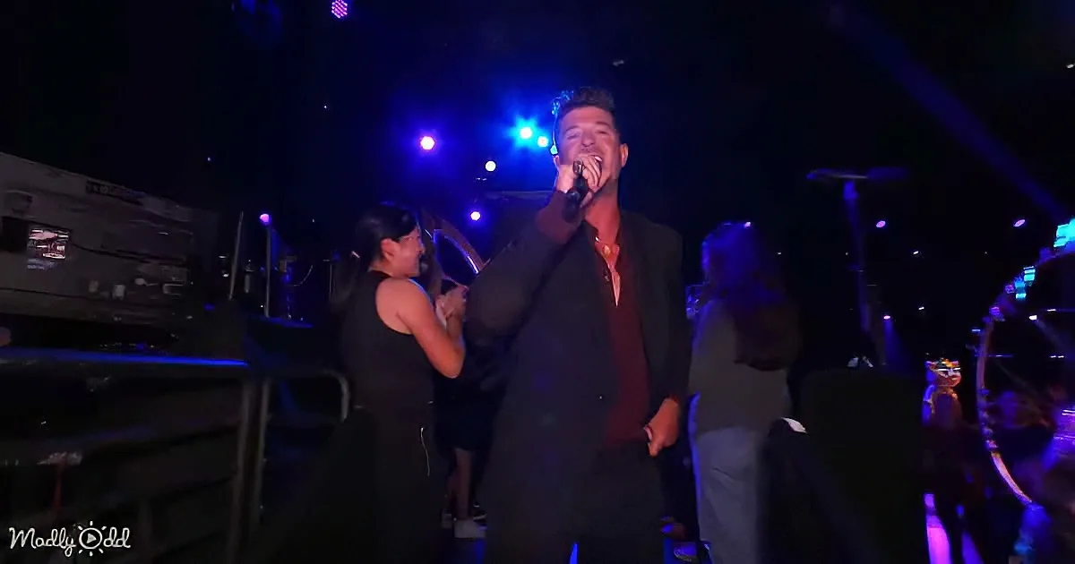 A man in a suit sings into a microphone on stage, surrounded by people and blue stage lights.