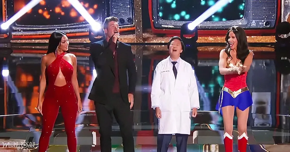 A man in a suit sings on stage with two women in red and a man in a lab coat.