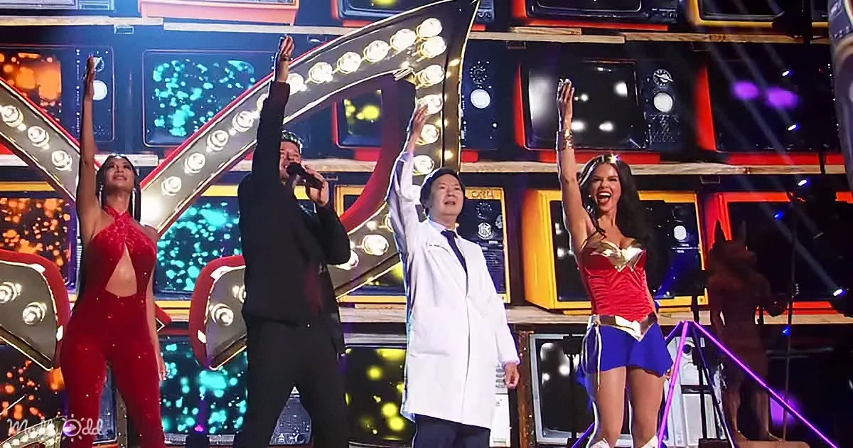 A man in a white lab coat raises his arm, surrounded by performers in costumes on a brightly lit stage.