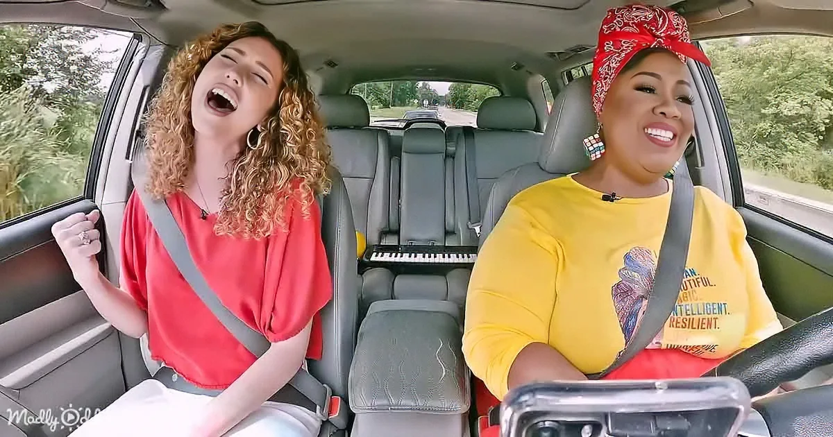 Two women in a car, one singing, the other smiling, with a keyboard in the back seat.