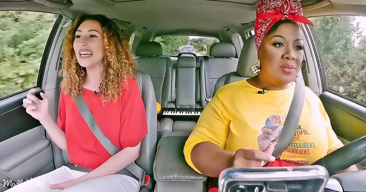Two women are driving and singing in a car, one in a red shirt and the other in a yellow shirt with a bandana.