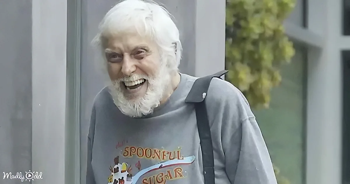 An elderly person with white hair and beard smiles widely, wearing a gray shirt with a colorful graphic.
