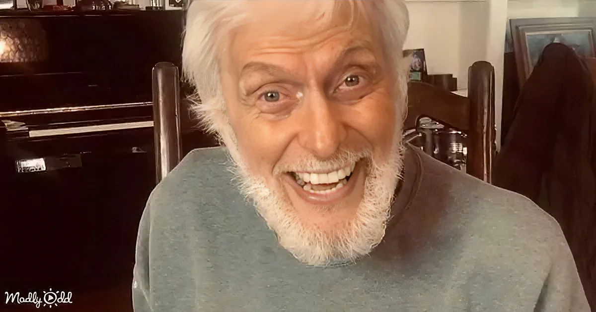 An older man with white hair and beard smiles widely, wearing a gray shirt, in a cozy indoor setting.