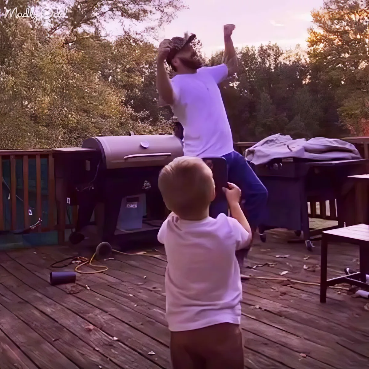 A man stands on a child's shoulders on a wooden deck, both wearing white shirts.
