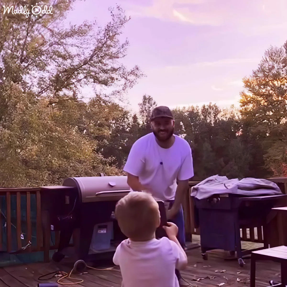 A man in a white shirt and cap stands on a deck, interacting with a child sitting on the ground.