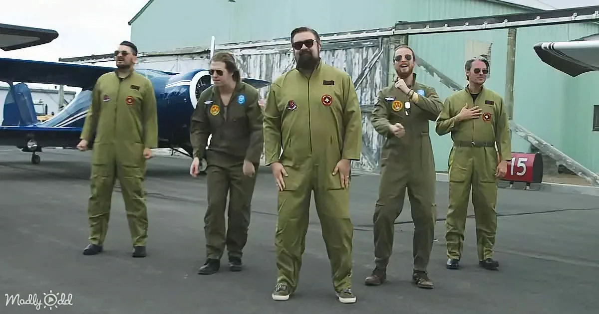 Five men in green flight suits stand on an airport tarmac, posing in front of a small aircraft.
