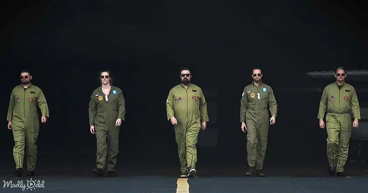 Five men in green flight suits walk in a line on a dark runway.