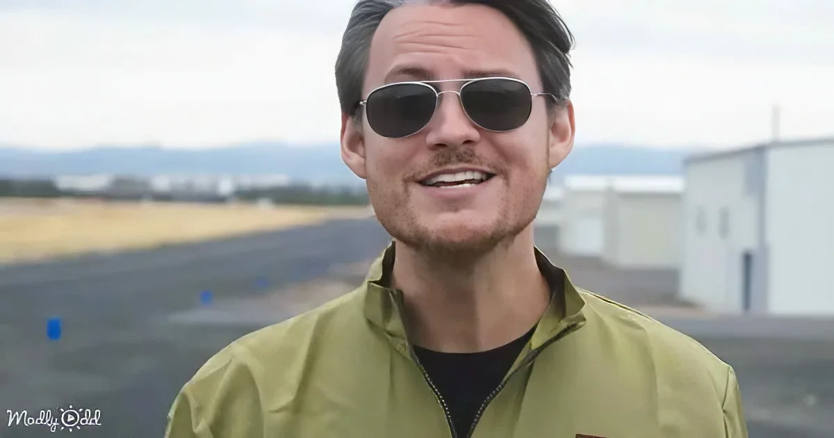 A man in a green jacket and sunglasses smiles, standing outdoors with a blurred background of buildings and a runway.